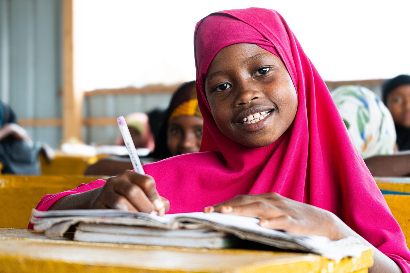 En flicka sitter vid en skolbänk i ett klassrum i Towfiq-lägret i Jowhar. Hon ler mot kameran medan hon skriver i sitt anteckningsblock. Ljuset faller in från sidan och andra elever syns i bakgrunden.