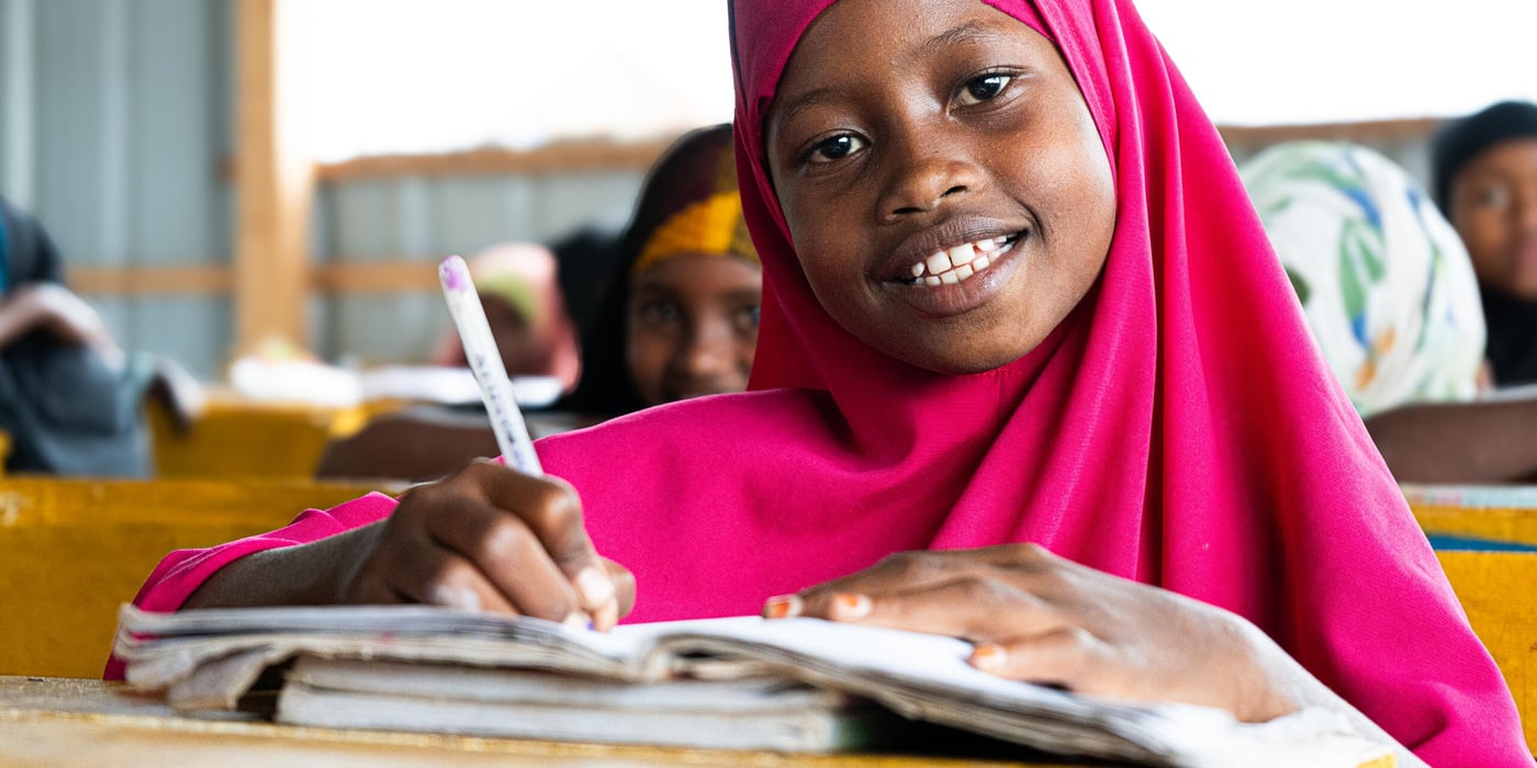 En flicka sitter vid en skolbänk i ett klassrum i Towfiq-lägret i Jowhar. Hon ler mot kameran medan hon skriver i sitt anteckningsblock. Ljuset faller in från sidan och andra elever syns i bakgrunden.