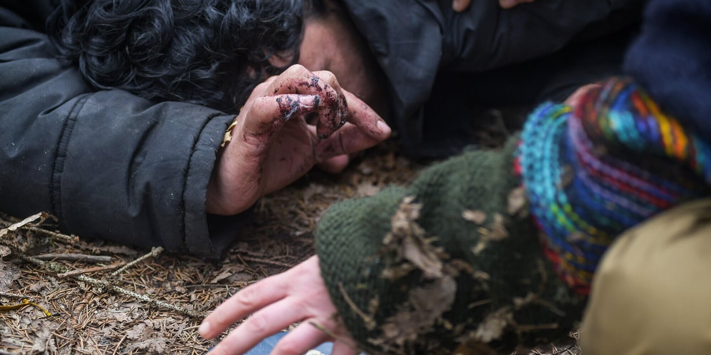 Person lying on the ground, with another person leaning over them to help. One of their hands is reaching for a passport.