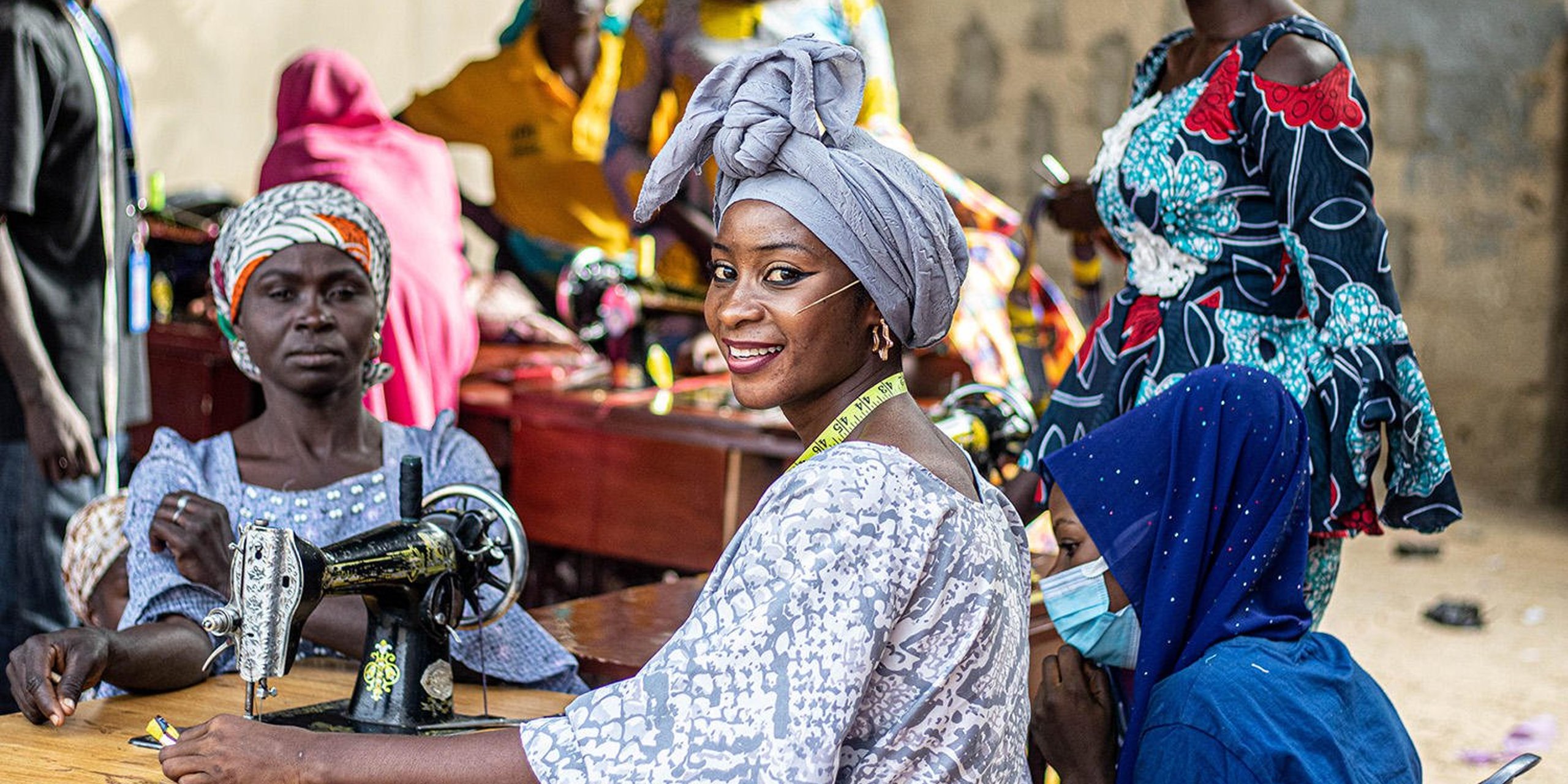 A woman is sitting by a sewing machine, teaching sewing to several other women. She is turned towards the camera, smiling.