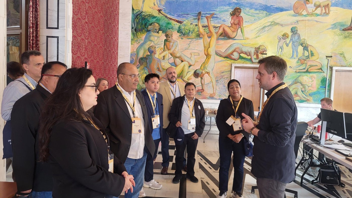 A group of men and women standing and listening to a man in the Oslo Town Hall building. Large mural painting on the wall behind them. 