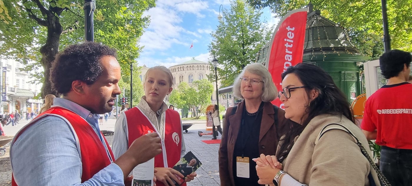 Two campaigners from the Norwegian Labour party, a young man and young woman, talking to two election observers on the streets of Oslo. Parliament in the background. 