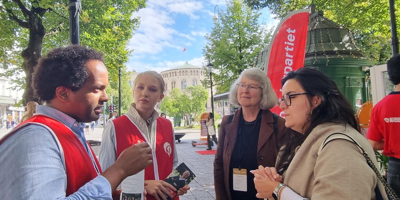 Two campaigners from the Norwegian Labour party, a young man and young woman, talking to two election observers on the streets of Oslo. Parliament in the background. 