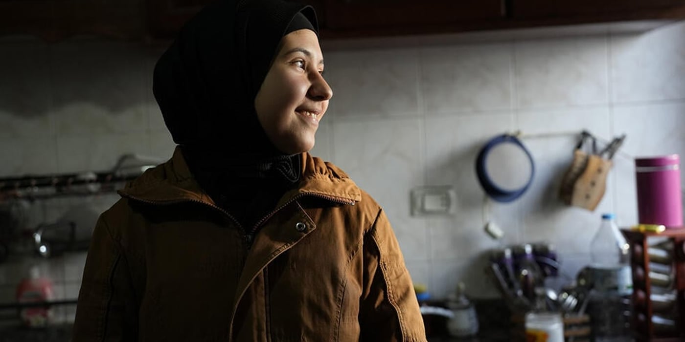 Young woman standing in a kitchen, smiling and looking at something to her left.