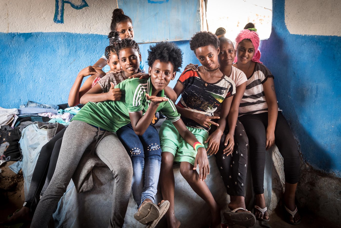 Winta, 16 years old, Furtuna, 15, Lidia, 14, Fiyori, 16, Fiyori, 14, Weyni, 15 and Adkonet, 12 live together in a house in Hitsat refugee camp just outside Shire, a district in northern Ethiopia. The camp houses about 10,000 refugees from Eritrea. Most of them are minors, and about one in ten have fled alone. 

The waiting room

Inside two small houses in a refugee camp in northern Ethiopia, seven girls and three boys wait for the Crown Prince Couple of Norway to visit.  All the children fled across the border from Eritrea, alone. “Off course we miss our families, but we cannot return now,” the children say.

It’s lunchtime and 15-year-old Furtuna sits next to a pot, stirring with all the strength she has. A sauce of tomatoes, paprika and onion simmer inside. She is preparing food together with the eldest children, while the youngest girls have other tasks, like fetching bread or cleaning. The seven girls, who live in the house together, didn’t know each other before they came to Hitsats refugee camp. 

“Now, we’re like sisters,” Winta says. For almost two years, the 16-year-old has stayed in the children’s collective run by the Norwegian Refugee Council (NRC). “Our friendship means a lot now.” 

The room is no larger than 20 square metres, yet the seven girls use it as a bedroom, a kitchen and a living room. They also share the three beds. On the left side of the house lives an adult who is responsible for the girls, watching out for them and helping them when needed. On the right side of the house live six boys who also fled Eritrea to Ethiopia, alone. 

As one of the eldest, Winta explains why they left their home country: “We’re all afraid to end up in the military and to lose the opportunity to get education. In the military you never finish and you have no choice. They come to get you, perhaps in the middle of the night. That’s why many of us had to leave.”

The girls crossed the border to Ethiopia on foot, all alone. “We heard people were shot on the way. In school, they told us that many girls were raped while trying to get away,” says Winta. The other girls nod their heads in affirmation. 

In the camp, the children attend school and get food. The girls are happy to live safely in the neighbouring country. “The very best is that we don’t have to be afraid anymore. And we have the opportunity to go to school and learn,” Winta says. 

The next door boys are 12, 14 and 15 years old. They have the same stories as the girls – grateful to be in safety, to escape the endless military service and to have the opportunity to go to school. 
“The education helps us grow and to make us stronger,” says 15-year-old Meyhaw. He came to the camp five months ago. 

When you ask the children if they have relatives in Europe, they all nod their heads. Uncles in Sweden and brothers in Germany. In the boys’ house, they plan to go further abroad. 

“We’re thinking that we have to travel to Sudan, Libya and then Europe. But for now it’s too strict, so in the meantime we’re happy to get more of an education,” says Meyhaw. 

The girls are not quite convinced as the boys about leaving Ethiopia. “I have a brother who left with a cousin, but only my brother made it over the sea,” says 15-year-old Weyni. “There are days when all I want is to travel, but then I feel it’s hopeless. We know how dangerous it is.”  

For now, the children want to stay in the children’s collective. The boys love their soccer practice and some of the girls have recently joined a drama group. If they were to wish for something better in life, they would wish for electricity to do homework. They would wish for some new clothes and maybe a cinema, so they could watch movies and forget about reality for a while. 

Photo: Beate Simarud/NRC 

Facts and figures:

Eritreans are the third largest group of refugees living in Ethiopia, with 37,321 refugees currently registered in refugee camps in the Shire area camps. 

Currently there are 163,281 Eritrean refugees in Ethiopia.

It is estimated that nearly 40 per cent of Eritrean refugees leave the camps within the first three months of arrival, and 80 per cent leave within the first year, with significant numbers of the population departing irregularly from Ethiopia to third countries – often with the assistance of smugglers and at great risk to their lives. 

The population in the Shire camps is unique, with a large number of children below the age of 18 and unaccompanied children. 

As of June 2017, 72 per cent of the refugees living in Shire were under the age of 18, including 4,725 unaccompanied and separated children, representing approximately 11.5 per cent of the total refugee population. 

Unaccompanied and separated children live in a variety of care arrangements, including community care, foster care, or family-based care.